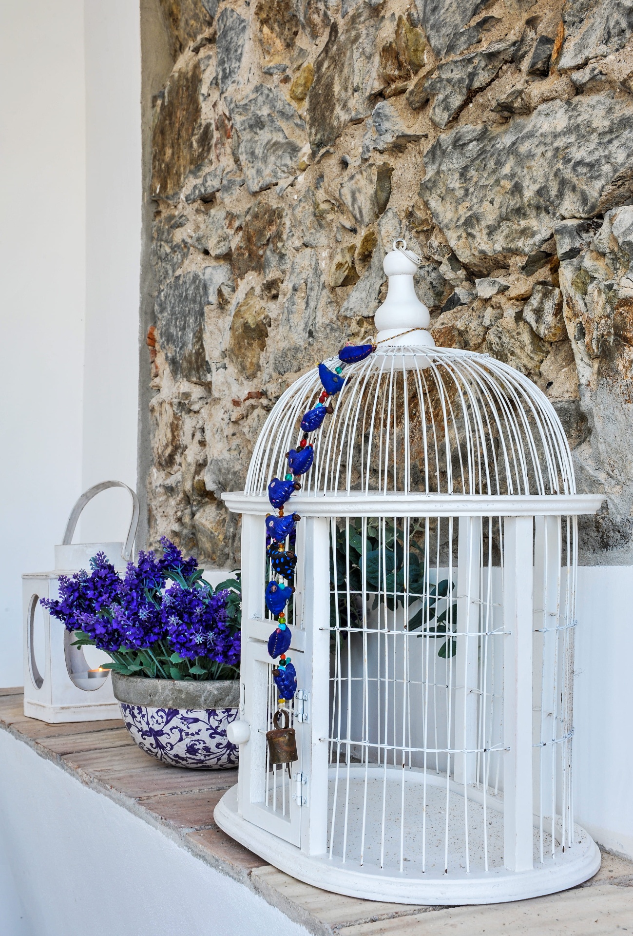 Detalhe decorativo com gaiola branca, flores roxas e parede em pedra